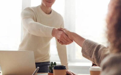 Two people shaking hands over a desk with a laptop and coffees