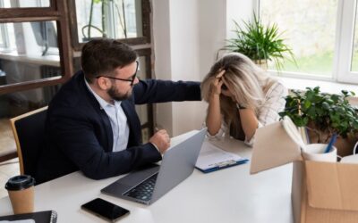 Man in a suit with a laptop in front of him resting his hand on the shoulder of a woman with her head in her hands