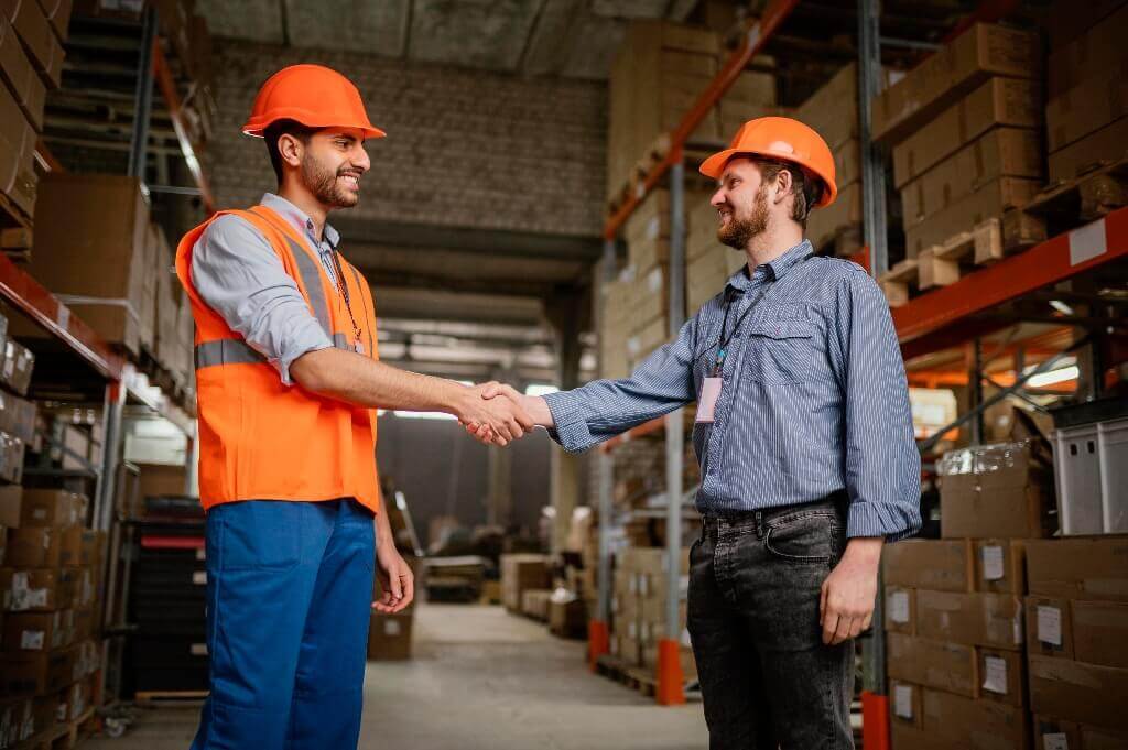Two men wearing hardhats shaking hands in a warehouse