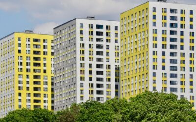 Modern high-rise apartment buildings with white and yellow facades, seen above a line of green trees under a cloudy sky.