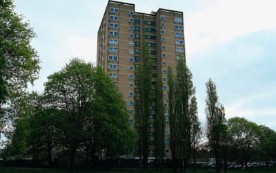 Tall residential apartment building surrounded by trees and greenery under an overcast sky.