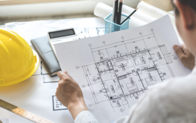 A person reviewing detailed architectural floor plans at a desk, with a yellow hard hat, ruler, notebook and calculator visible nearby.
