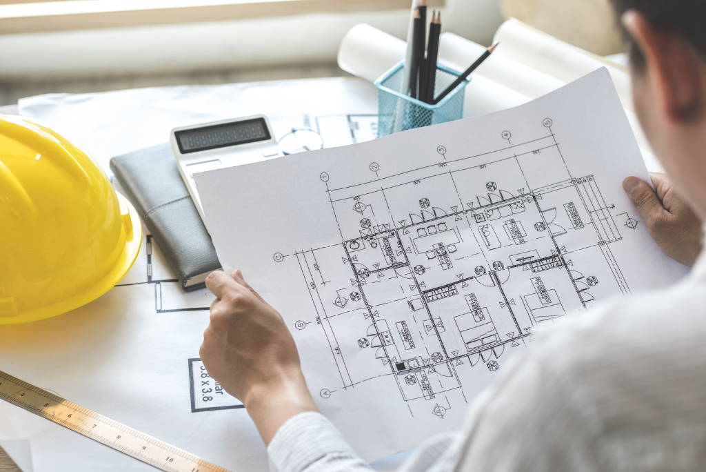A person reviewing detailed architectural floor plans at a desk, with a yellow hard hat, ruler, notebook and calculator visible nearby.