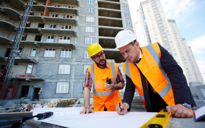 Two construction professionals in high-visibility orange vests and hard hats reviewing building plans on a surface outdoors, with a multi-storey building under construction visible in the background.