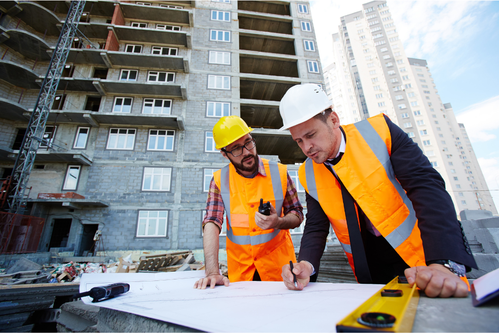 Two construction professionals in high-visibility orange vests and hard hats reviewing building plans on a surface outdoors, with a multi-storey building under construction visible in the background.
