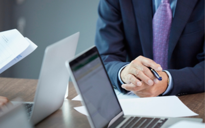A business executive in a suit sits at a meeting table with hands clasped around a pen, surrounded by laptops, financial documents and reports, suggesting a discussion about corporate governance, financial performance and insolvency-related decision-making.
