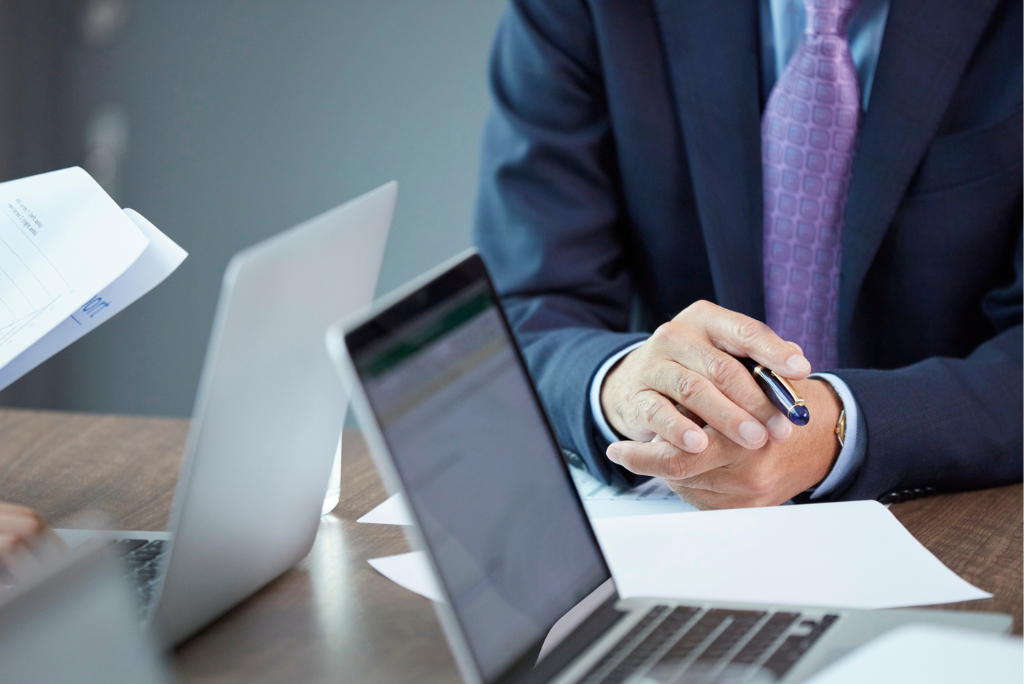 A business executive in a suit sits at a meeting table with hands clasped around a pen, surrounded by laptops, financial documents and reports, suggesting a discussion about corporate governance, financial performance and insolvency-related decision-making.