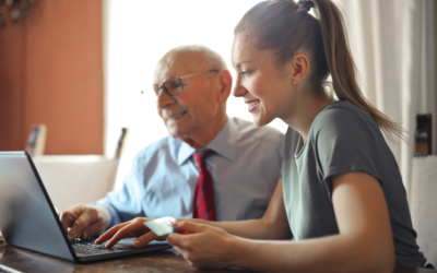 Young woman helping an older man use a laptop at a table in a bright home. Both are smiling as they look at the screen, suggesting support financial planning.