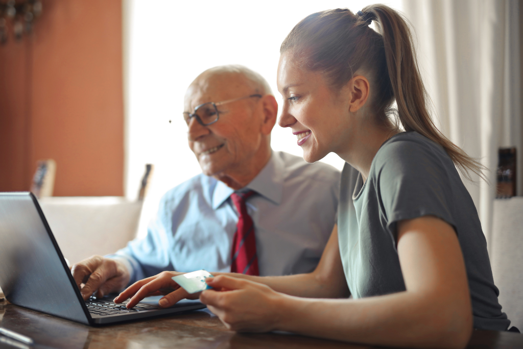 Young woman helping an older man use a laptop at a table in a bright home. Both are smiling as they look at the screen, suggesting support financial planning.