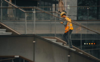 Construction worker wearing a hard hat and high-visibility vest walks up a modern outdoor staircase while looking at a mobile phone.