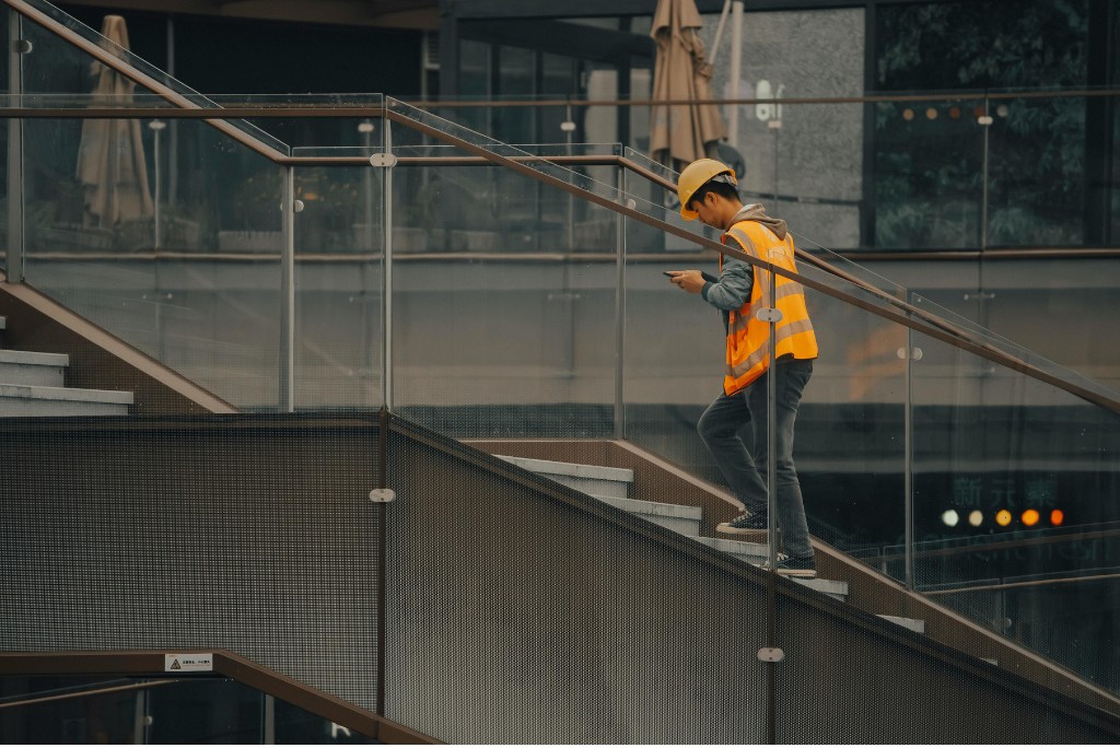 Construction worker wearing a hard hat and high-visibility vest walks up a modern outdoor staircase while looking at a mobile phone.