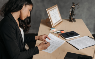 A professional woman in business attire sits at a desk reviewing and signing legal documents, with a laptop, notebook, clipboard, and a statue of Lady Justice visible in the background, creating a formal legal or financial advisory setting.