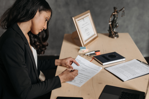 A professional woman in business attire sits at a desk reviewing and signing legal documents, with a laptop, notebook, clipboard, and a statue of Lady Justice visible in the background, creating a formal legal or financial advisory setting.