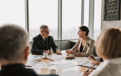 Business professionals gathered around a meeting table discussing documents and reports in a modern office. Two colleagues lead the conversation while others take notes, with large windows overlooking the city and financial charts spread across the table.