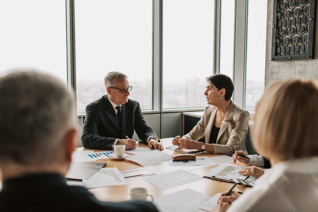 Business professionals gathered around a meeting table discussing documents and reports in a modern office. Two colleagues lead the conversation while others take notes, with large windows overlooking the city and financial charts spread across the table.