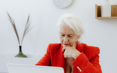 Older woman in a red jacket sits thoughtfully at a laptop in a bright home office, resting her chin on her hand while looking at the screen. The image conveys careful consideration of financial, pension or legal matters in a calm, modern setting.