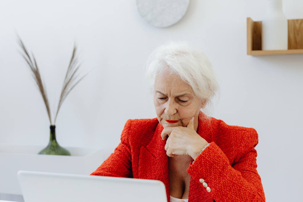 Older woman in a red jacket sits thoughtfully at a laptop in a bright home office, resting her chin on her hand while looking at the screen. The image conveys careful consideration of financial, pension or legal matters in a calm, modern setting.