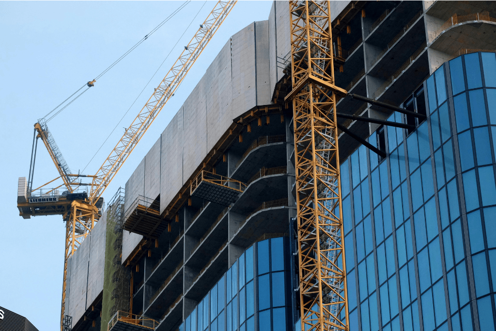 Construction site with two large cranes working on a modern multi-story building featuring glass windows and exposed concrete structure.